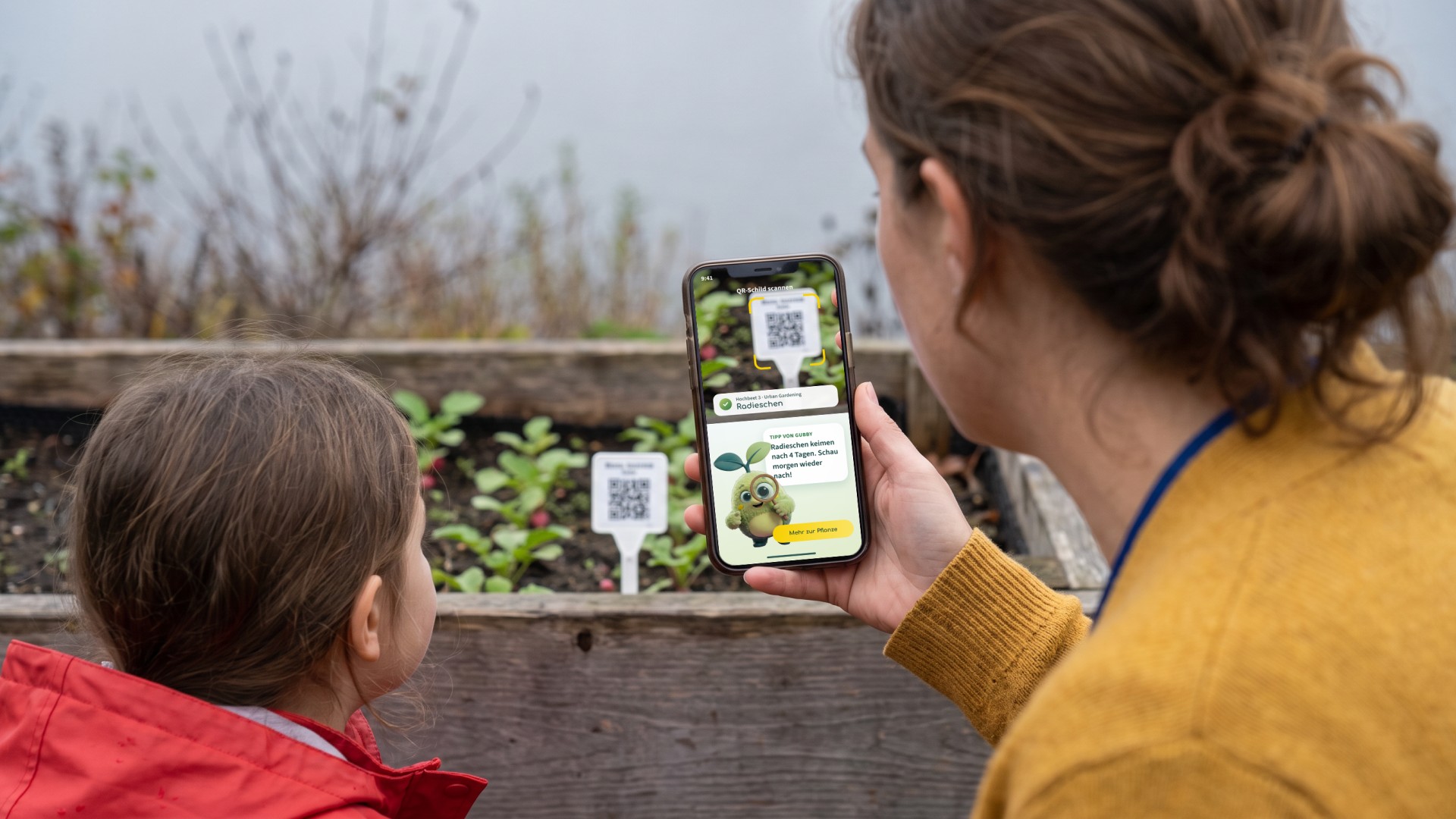 Blick über die Schulter einer Erzieherin am Hochbeet: Sie scannt mit dem Handy das QR-Schild bei den Radieschen, ein Mädchen schaut zu. Auf dem Handy erkennt die App das Beet, darunter gibt Gubby mit der Lupe einen Tipp.